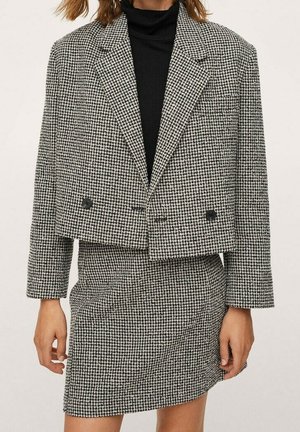 Kobieta ubrana w czarny golf pod czarno-białą krótka marynarkę w wzór pepitki i dopasowaną spódnicę, stojąca na jednolitym tle.