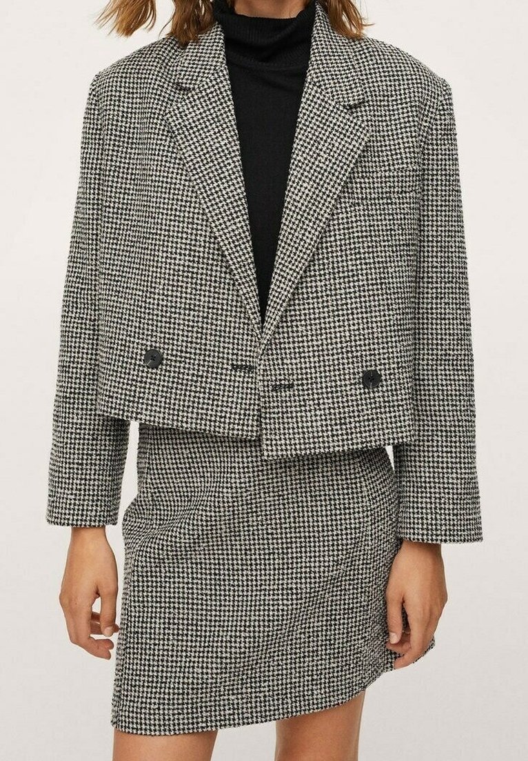 Femme portant un col roulé noir sous un blazer court pied-de-poule noir et blanc et une jupe assortie, debout devant un fond uni.