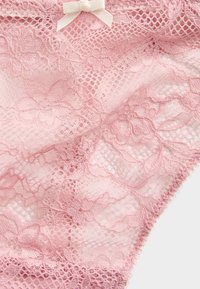 Lingerie di pizzo rosa con motivo floreale, caratterizzata da un delicato motivo a fiocco in vita. Materiale testurizzato e semi-trasparente.