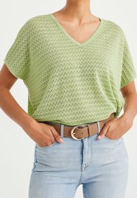 Vaaleanvihreä V-pääntie paita, jossa on teksturoitu kuvio, yhdistettynä korkeavyötäröisiin vaaleansinihiin farkkuihin ja ruskeaan vyöhön, jossa on pyöreä metallisolki.