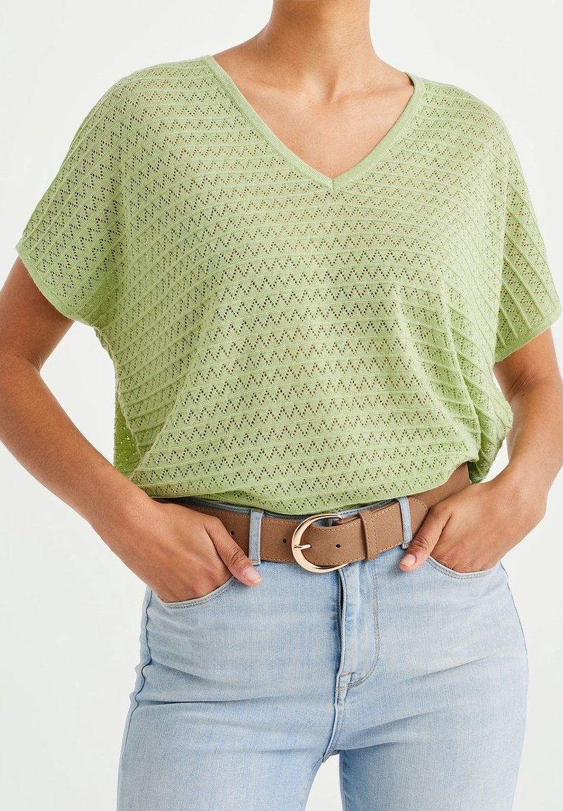 Vaaleanvihreä V-pääntie paita, jossa on teksturoitu kuvio, yhdistettynä korkeavyötäröisiin vaaleansinihiin farkkuihin ja ruskeaan vyöhön, jossa on pyöreä metallisolki.