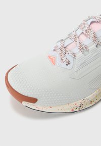 Baskets de sport gris clair avec une tige texturée, des lacets à motifs et un capuchon en cuir marron. La semelle présente un design moucheté dans plusieurs couleurs.