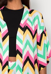 Multikleurige lange kimono met een zigzagpatroon in roze, geel, groen, zwart en wit. Gemaakt van lichtgewicht stof, heeft korte mouwen.