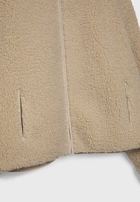 Beżowa kurtka z polaru o miękkiej, teksturowanej powierzchni; posiada boczne rozcięcia i gładką podszewkę wzdłuż zamka.