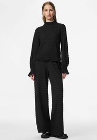 Crni teksturirani dvojezični outfit koji se sastoji od dugih rukava s visokim ovratnikom i naboranim manžetama, uparen s hlačama širokih nogavica.