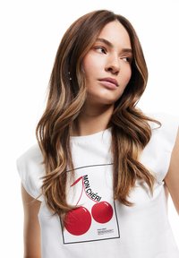 Camisa de manga corta blanca con un estampado gráfico de cerezas rojas y el texto "MON CHÉRI", con una textura suave y ajuste regular.