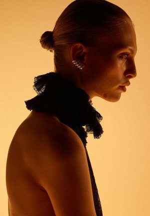 Black lace collar with ruffled edges, metallic earring, smooth skin, and a bun hairstyle against a warm orange background.