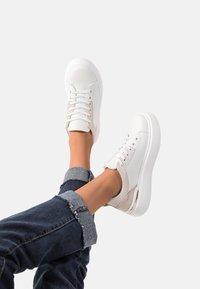 Sneaker bianchi con design a lacci, materiale in tela e un accento beige chiaro. Punta arrotondata e suola spessa. Jeans con orli risvoltati.
