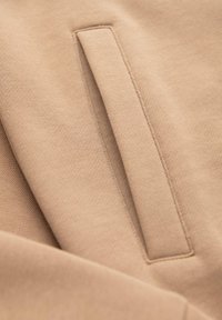 Tissu beige avec une poche passepoilée rectangulaire cousue dans le matériau, montrant la texture et les coutures en gros plan.