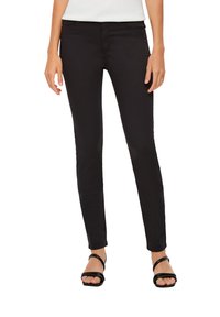 Jean skinny noir avec une texture de tissu lisse, présentant un design classique à cinq poches et une taille mi-haute. Porté avec des sandales noires.