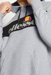 Sweat à capuche gris avec une bande horizontale noire et un grand logo "ellesse". Cordon de serrage noir et poche avant visibles. Tissu texturé.