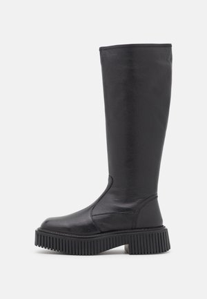 Botte haute en cuir noir avec semelle crantée épaisse et bout arrondi, conçue pour la mode et la durabilité.