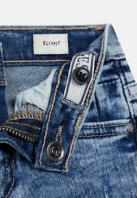 Primo piano di jeans slim-fit blu in denim sbottonati che mostrano il etichetta in vita, bottone, zip e dettagli delle cuciture.