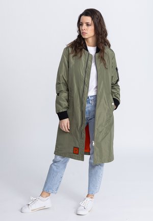 Olijfgroene lange jas met een ritssluiting en oranje voering, met een patch aan de voorkant en zwarte gebreide manchetten. Gemaakt over denim en sneakers.