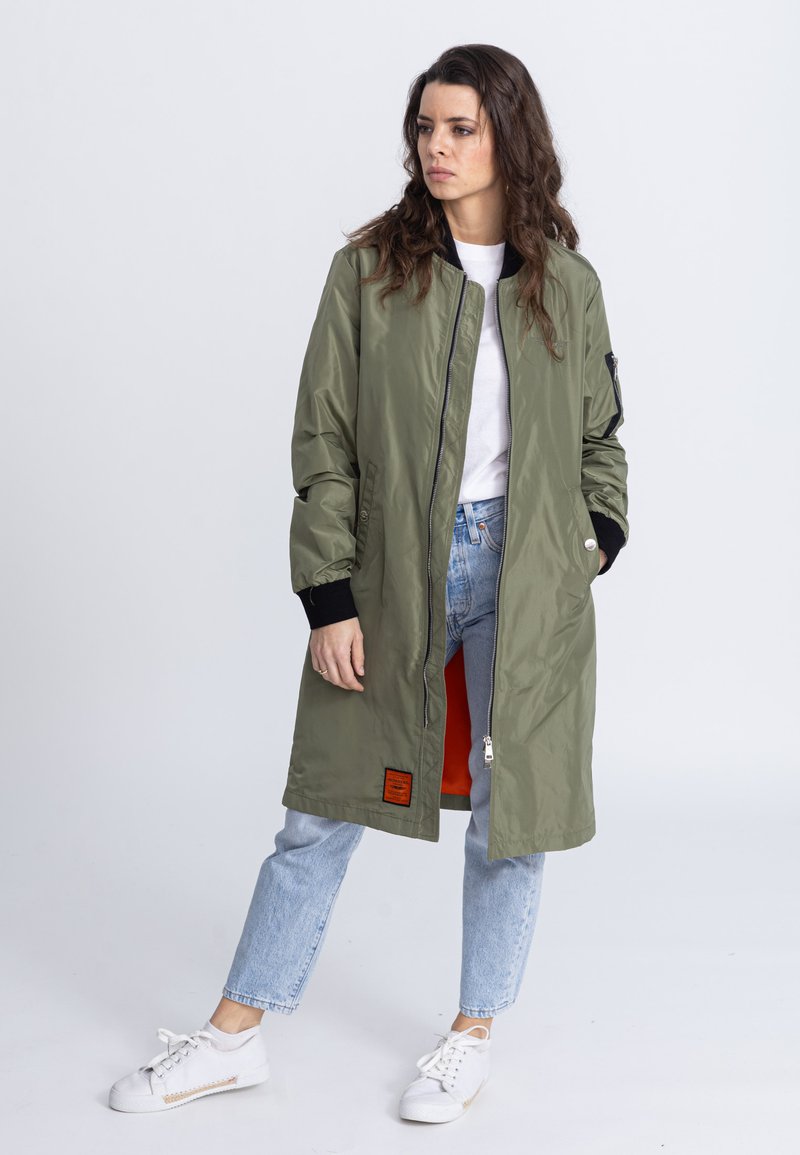 Olivenfarbener langer Mantel mit Reißverschluss und orangefarbenem Futter, ausgestattet mit einem Patch auf der Vorderseite und schwarzen Strickbündchen. Getragen über Jeans und Sneakern.