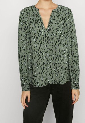 Femme portant un chemisier vert à manches longues avec des taches abstraites noires et blanches, assorti à un pantalon noir sur un fond uni.