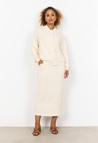 Outfit color crema composto da una maglia a maniche lunghe con zip e una gonna midi coordinata con tasche, realizzato in un tessuto morbido e testurizzato.