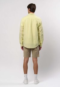 Mann mit blassgelbem Langarmshirt, beigen Shorts, weißen Socken und weißen Turnschuhen, stehend vor einem schlichten hellgrauen Hintergrund.