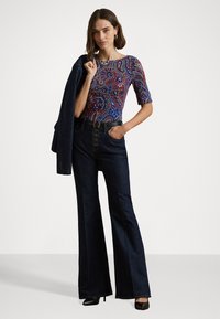 Jeans in denim scuro a gamba svasata con vita alta e chiusura a bottoni. Top aderente a fantasia in rosso, blu e bianco, abbinato a una giacca scura.
