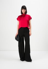 Femme debout contre un fond blanc, portant un top en satin rouge, un pantalon large noir, tenant une pochette noire.