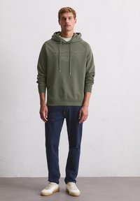 Hoodie vert olive avec cordons de serrage, logo détaillé, poignets côtelés, associé à un jean bleu foncé et des baskets blanches. Texture lisse dans l'ensemble.