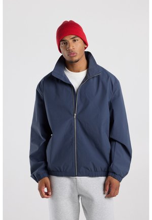 Jonge man met een rode muts, marineblauwe jas met rits, wit overhemd en lichtgrijze joggingbroek tegen een effen lichte achtergrond.
