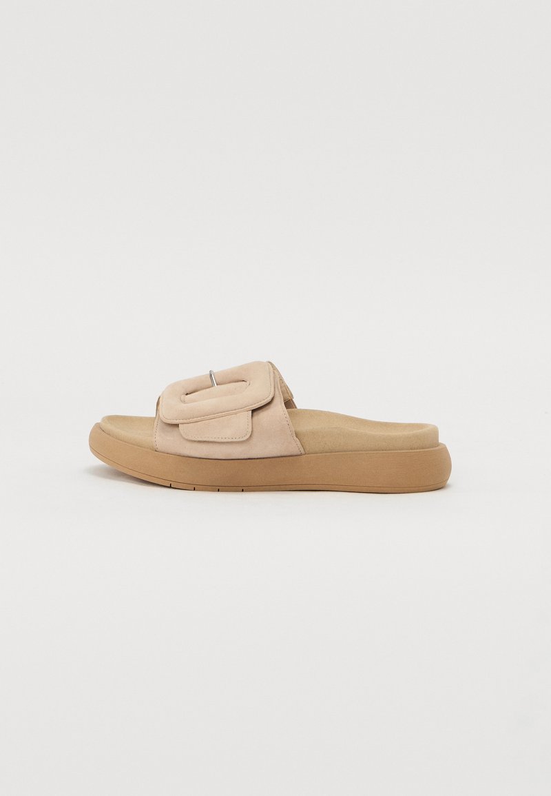 Beige suede slide sandal with a cushioned footbed, rounded toe, slightly elevated sole, and a decorative strap featuring a silver accent.