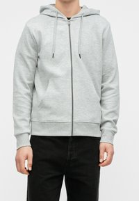 Grauer Kapuzenpullover mit Reißverschluss aus weichem Stoff, mit Fronttaschen, Kordelzugkapuze, gerippten Bündchen und Saum, kombiniert mit schwarzer Hose.