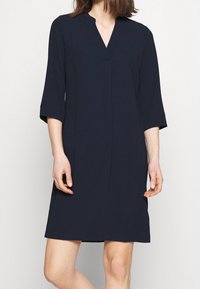 Marinblå klänning med V-krage, trekvartslånga ärmar och rak silhuett. Tillverkad av ett slätt tyg med minimal detaljer.