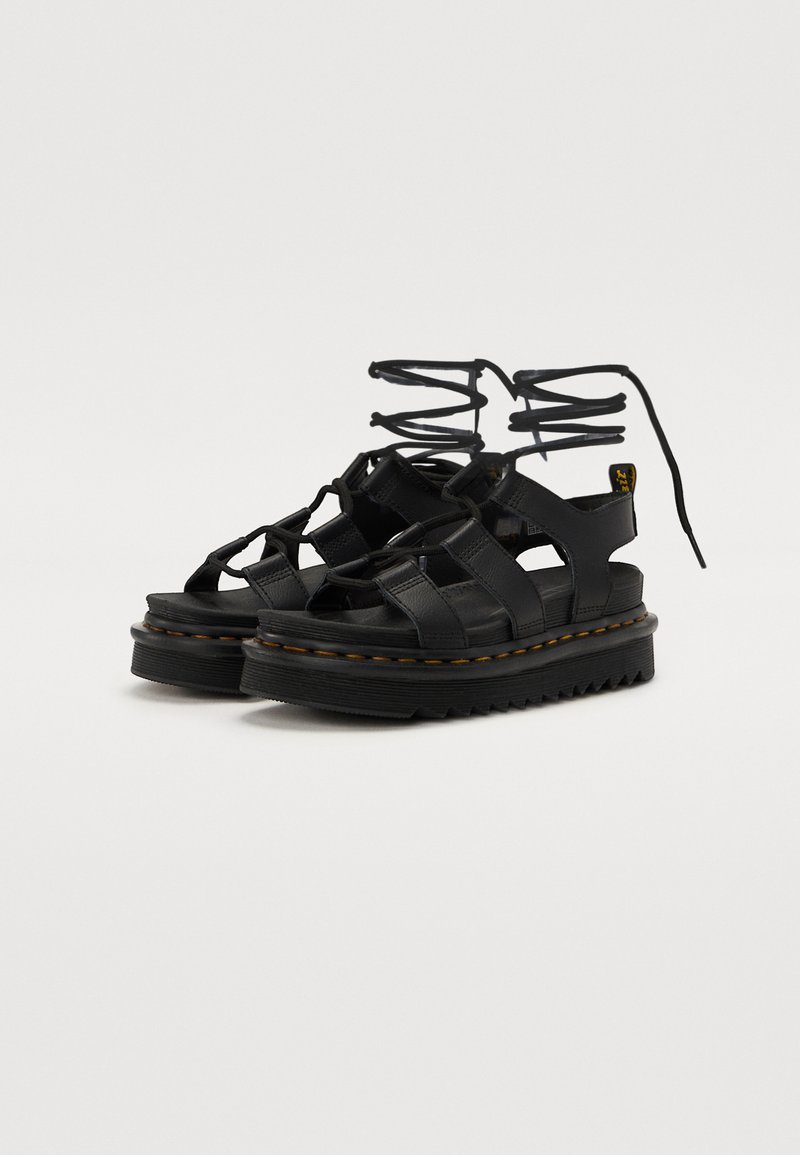 Black leather platform sandals with strap design, featuring a thick rubber sole and visible yellow stitching accents. Laces extend from the ankle.
