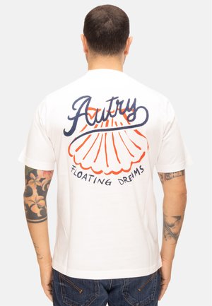 Hombre con brazos tatuados lleva camiseta blanca con concha naranja y texto azul "Autry Floating Dreams" en la espalda, de espaldas.
