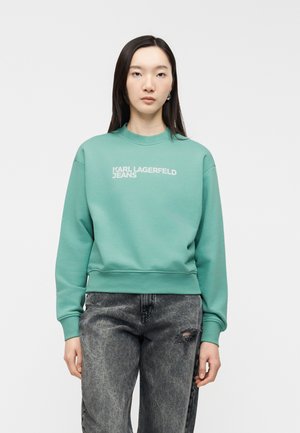 Nő türkiz színű pulóverben, amelyen a "KARL LAGERFELD JEANS" felirat szerepel, fekete savas mosású farmerral, amelyen kis szakadás van a combnál.