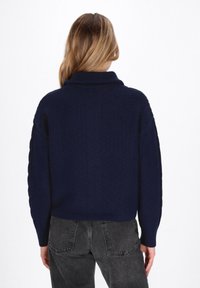 Marineblauer Strickpullover mit hohem Kragen, texturiertem Muster und überschnittenen Schultern. Kombiniert mit hochtaillierten dunklen Jeans, von hinten betrachtet.