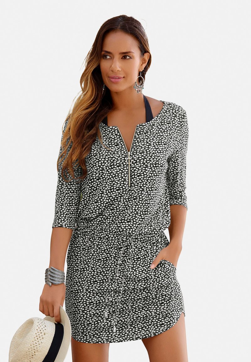 Schwarz-weiß gemustertes Kleid mit drei Viertelärmeln, einem Reißverschluss am Hals, taillierter Taille und Seitentaschen; das Model hält einen Strohhut.