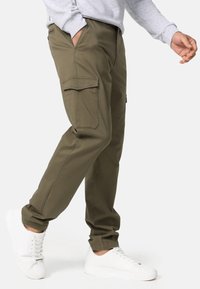Pantalones cargo verde oliva con bolsillos laterales, diseño de pierna ajustada, material de algodón, combinados con zapatillas blancas. La textura parece suave.