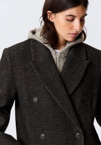 Dark houndstooth double-breasted coat featuring large black buttons and a notched lapel, layered over a light gray zip-up hoodie.