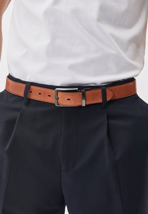 Ceinture en cuir marron avec une boucle en argent rectangulaire, portée sur un pantalon marine et une chemise blanche. Présente plusieurs trous pour l'ajustement.