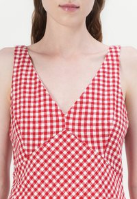 Punainen ja valkoinen ruudullinen mekko, jossa on V-pääntie, hihaton muotoilu ja napakka yläosa, jossa on kolmiomainen sauma yksityiskohtia. Puuvillamateriaali.