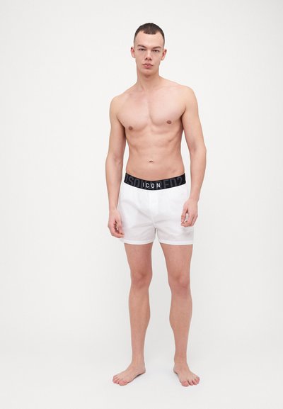Boxer shorts blancs avec une taille noire comportant le mot "ICON" dans un motif en répétition. Tissu lisse, coupe droite, longueur au-dessus du genou.