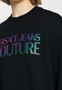 Svart sweatshirt med en iriserande logotyp som säger "VERSACE JEANS COUTURE," med en klassisk rund halsringning och textilstruktur.