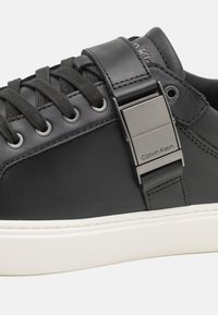 Baskets en cuir noir avec un design épuré, lacets texturés, boucle en métal argenté et semelle en caoutchouc blanc. Logo discrètement affiché.