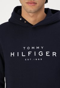 Námořnická modrá mikina z měkké tkaniny s bílým tištěným logem značky "TOMMY HILFIGER" a rokem založení "EST.1985" na přední straně.