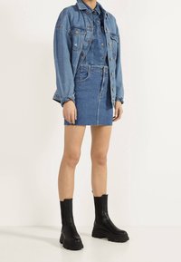 Kvinna iklädd en blå denimjacka, matchande denimkjol med rå fåll och svarta kraftiga stövlar som når till vaden, stående mot en enkel vit bakgrund.