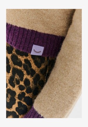 Dzianinowy sweter w beżowym kolorze z fioletowym prążkowanym wykończeniem na dole i akcentami w panterkę. Zawiera małą lawendową metkę z graficznym detalem.