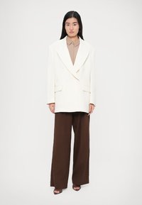 Biely dvojradový blejzer so širokými klopami, nosený cez béžovú košeľu na gombíky, v kombinácii s voľnými hnedými nohavicami a sandálmi s odkrytými prstami.