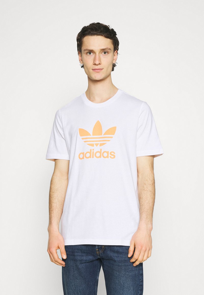 T-shirt bianco in cotone con il logo trefoil arancione di Adidas sul petto. Maniche corte e colletto rotondo, vestibilità rilassata.