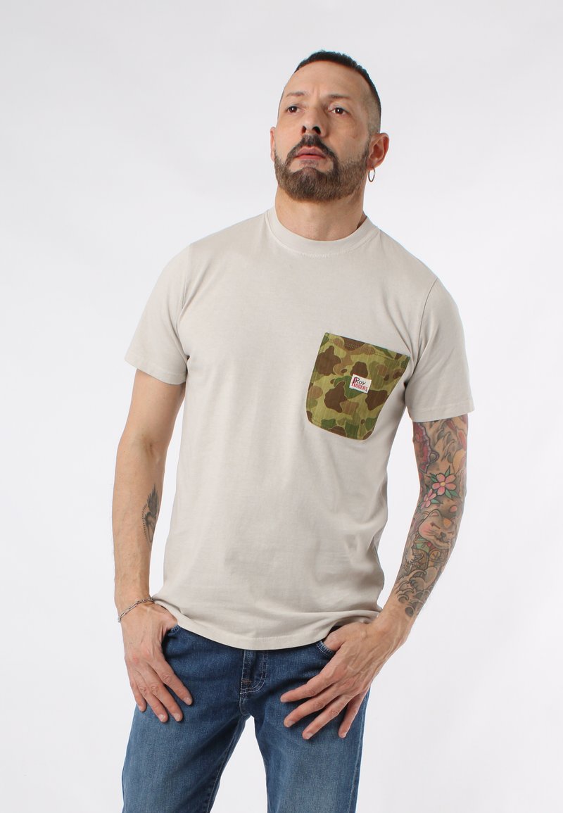 Maglietta di cotone beige con taschino camouflage e etichetta del marchio. Maniche corte, collo rotondo, vestibilità rilassata, abbinata a jeans blu.