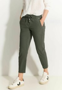 Olivengroene tapered broek met een elastische tailleband, trekkoord, zijzakken en een gladde textuur, gecombineerd met witte sneakers.