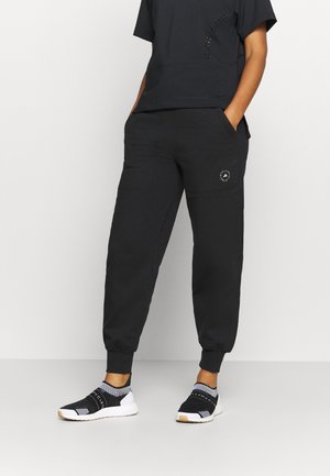Person wearing black jogger pants with logo on thigh, black top, and black-and-white slip-on athletic shoes, standing with hands in pockets.