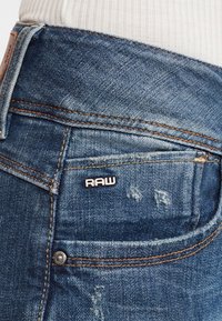 Närbild av en blå denimjeansficka med orange sömnad och en liten svart och vit "RAW"-etikett.